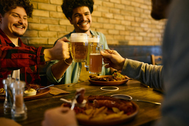 Κοντινό πλάνο χαρούμενων αρσενικών φίλων που κάνουν πρόποση με μπύρα ενώ μαζεύονται για μεσημεριανό σε μια παμπ.  - Φωτογραφία, εικόνα