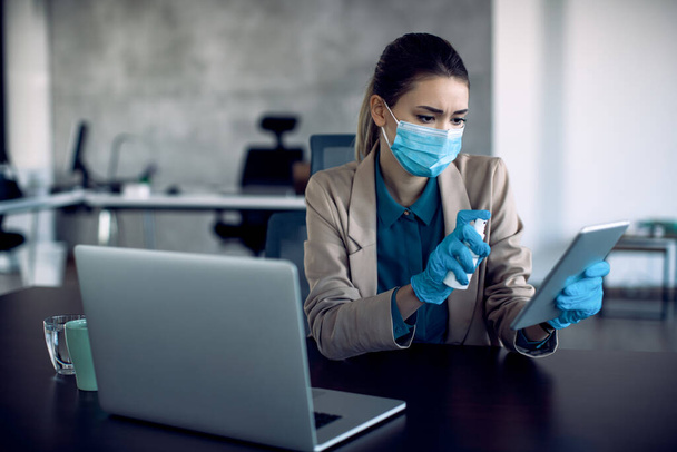 Businesswoman cleaning touchpad with disinfectant while wearing face mask and protective gloves in the office due to COVID-19 pandemic.  - Photo, image