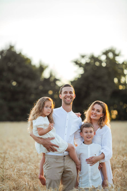 Beautiful young family at sunset in a wheat field. Happy parents with two children, a boy and a girl, in light clothes. Summer. Love. - Foto, afbeelding