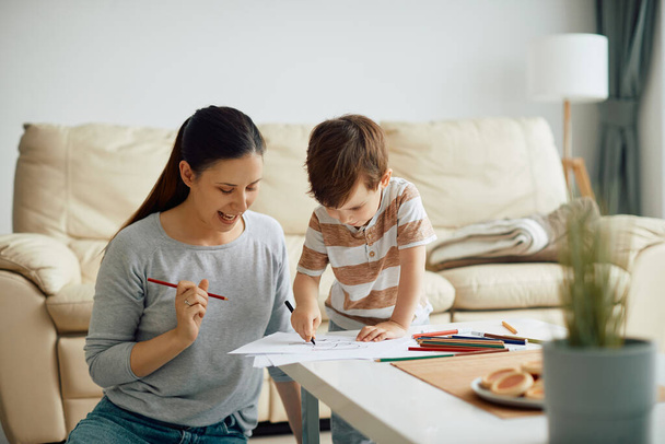 Young mother having fun and drawing on the paper with her son at home. - 写真・画像