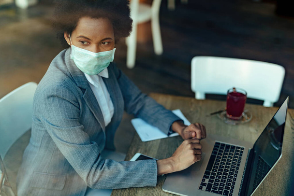 Yüzünde koruyucu maskeyle bir kafede otururken dizüstü bilgisayar kullanan dalgın siyah bir iş kadını. - Fotoğraf, Görsel