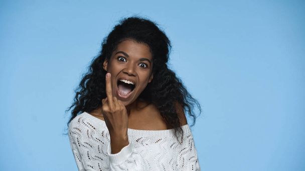happy african american woman in white knitted sweater showing middle finger isolated on blue - Φωτογραφία, εικόνα