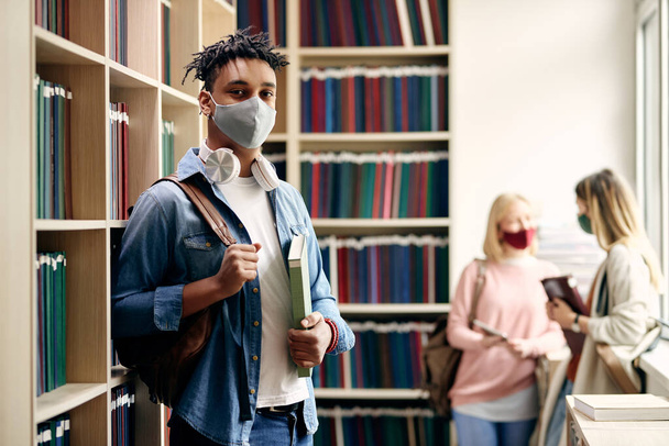 Portrait of smiling black student wearing protective face mask while studying in library during coronavirus pandemic. There are students in the background.  - Фото, зображення