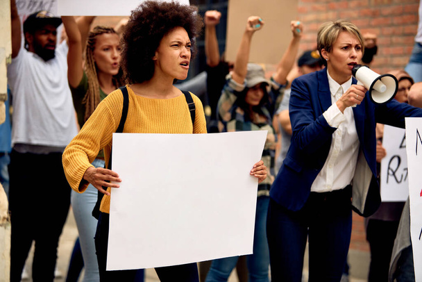Велика група незадоволених демонстрантів марширують на демонстрації по вулицях. Зосереджений на афро-американській жінці, яка тримає чистий плакат. Копіювальний простір. - Фото, зображення