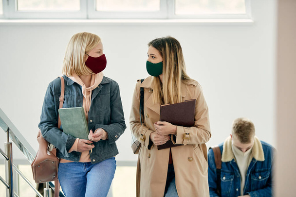 Happy female friends communicating while wearing protective face masks and walking through university hallway.  - Foto, afbeelding