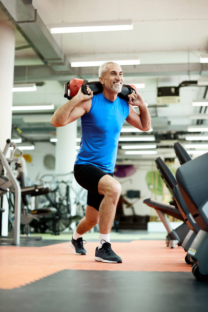 Happy mature athlete in lunge position doing sandbag shouldering exercises during sports training in a gym.  - Foto, Imagem