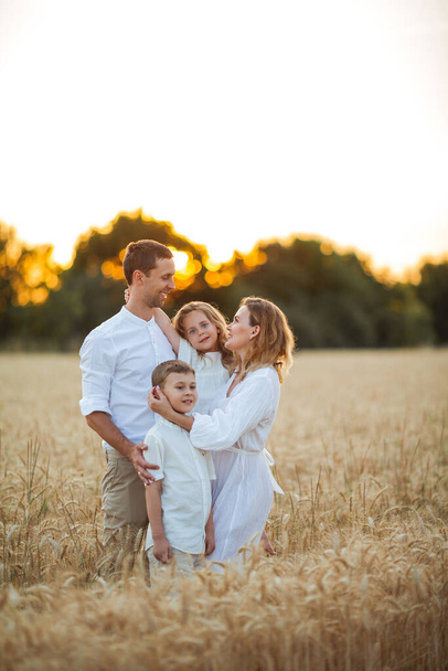 Beautiful young family at sunset in a wheat field. Happy parents with two children, a boy and a girl, in light clothes. Summer. Love. - Fotó, kép