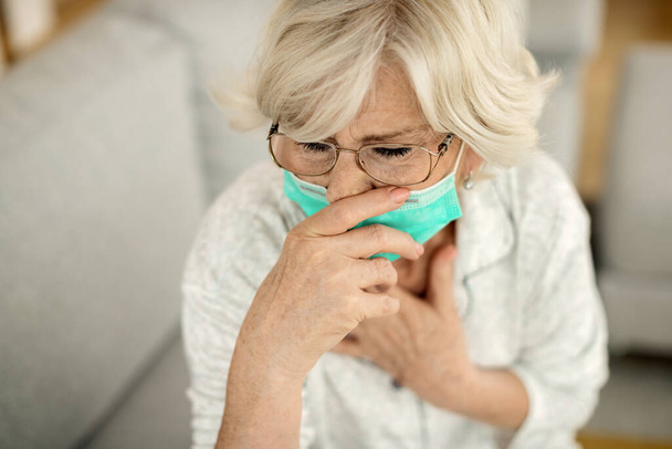 Ηλικιωμένη γυναίκα με μάσκα προσώπου που αισθάνεται άρρωστος και βήχει στο σπίτι.  - Φωτογραφία, εικόνα