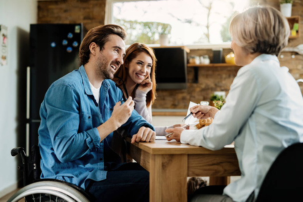 Szczęśliwy mężczyzna na wózku inwalidzkim i jego żona za pomocą tabletu cyfrowego z doradcą finansowym podczas spotkania w domu.  - Zdjęcie, obraz