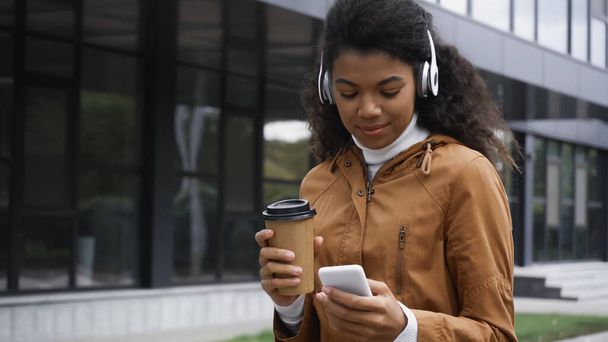 göndör afro-amerikai nő vezeték nélküli fejhallgató gazdaság okostelefon és papír pohár kívül  - Fotó, kép