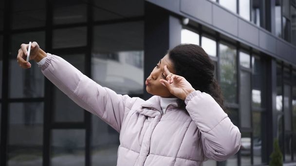 curly african american woman in puffer jacket taking selfie on smartphone and showing peace sign outside - Photo, image