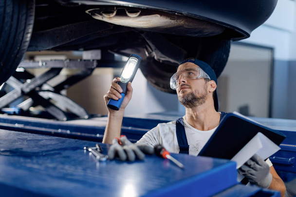 Mid adult repairman using flashlight while examining undercarriage of a car in auto repair shop. - Foto, Imagem