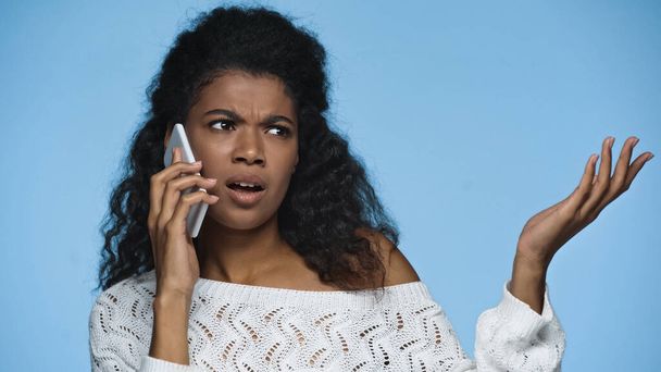 displeased african american woman in knitted sweater gesturing while talking on cellphone isolated on blue - Foto, Imagem