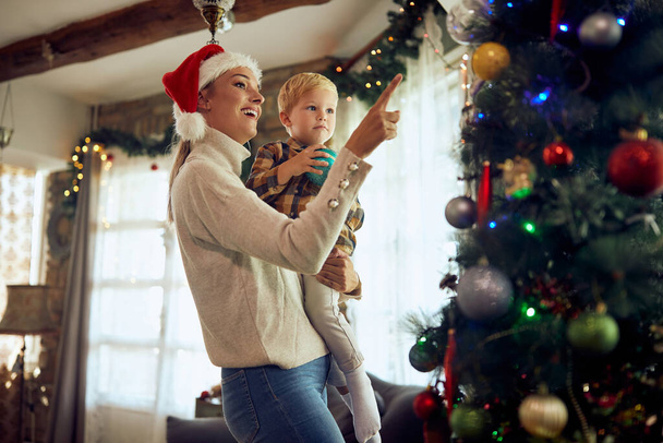 Ευτυχισμένη μητέρα και γιος διασκεδάζουν ενώ στολίζουν το χριστουγεννιάτικο δέντρο στο σαλόνι. - Φωτογραφία, εικόνα