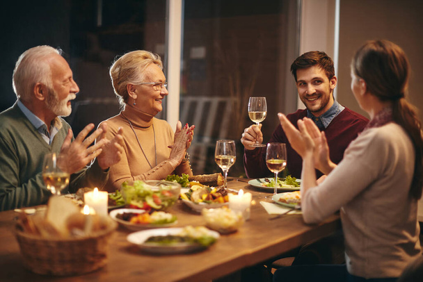 Щасливий чоловік виховує келих, поки його сім'я аплодує під час обіду за обіднім столом. Фокус на старшій жінці
.  - Фото, зображення