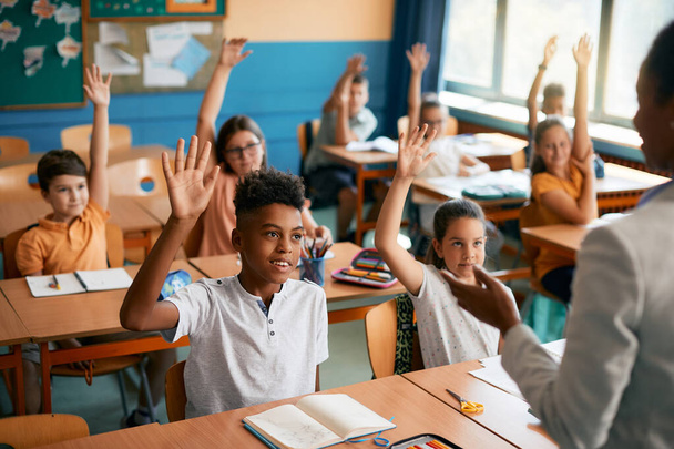 Πολυεθνική ομάδα ευτυχισμένων μαθητών που σηκώνουν τα χέρια για να απαντήσουν στην ερώτηση του δασκάλου για μια τάξη στην τάξη. - Φωτογραφία, εικόνα
