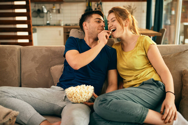 Happy woman having fun while boyfriend is feeding her with popcorn in the living room.  - Zdjęcie, obraz