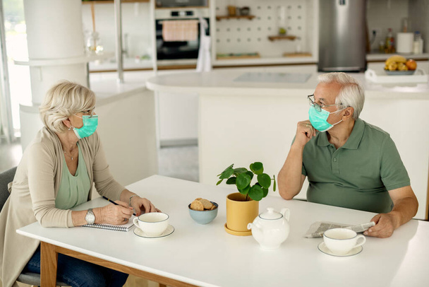 Mature man reading newspaper and communicating with his wife at home. Both of them are wearing protective face masks due to coronavirus epidemic.  - Foto, imagen