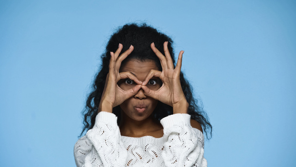 african american woman in knitted sweater grimacing and showing hand gesture symbolizing binoculars isolated on blue - Fotoğraf, Görsel