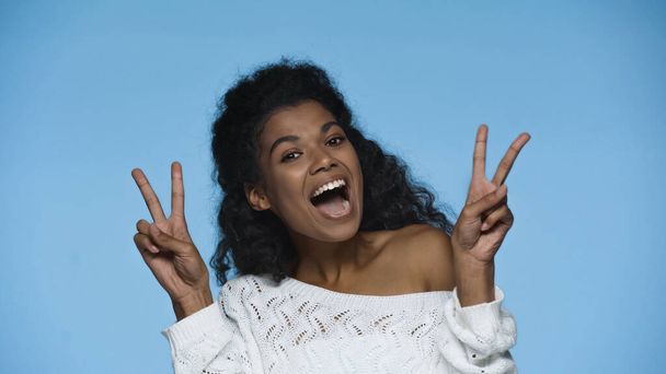 amazed african american woman in white knitted sweater showing peace sign isolated on blue - Foto, Bild