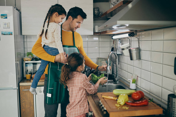 Середній дорослий батько і його маленькі дочки миють овочі під час коксування на кухні
.  - Фото, зображення