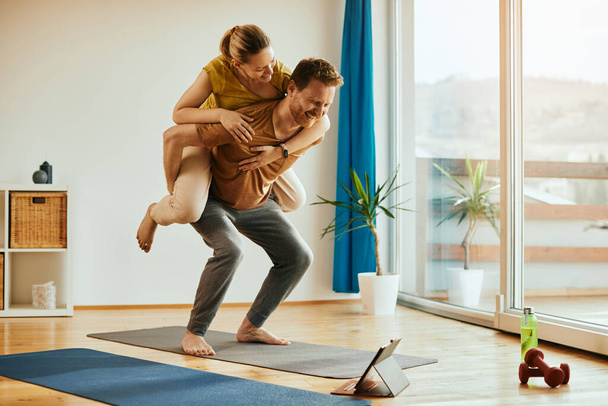 Happy man piggybacking his girlfriend while practicing squats in the living room.  - Valokuva, kuva