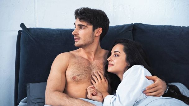 shirtless boyfriend and smiling girlfriend in white shirt looking away while lying on bed - Фото, изображение