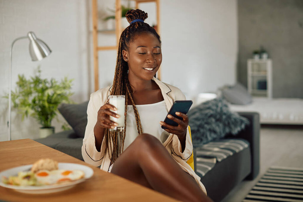 Smiling black woman having a glass of milk and using smart phone during her breakfast at home. - Zdjęcie, obraz