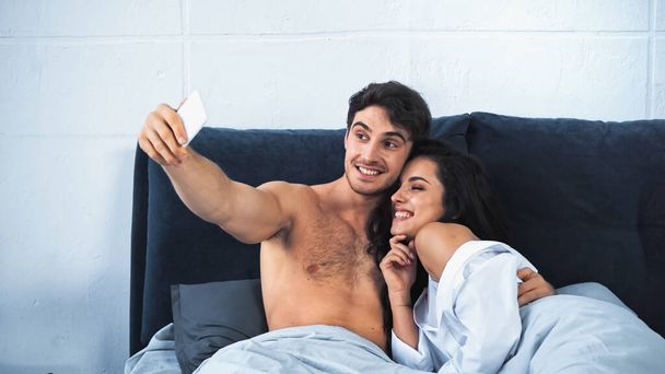 happy and shirtless man taking selfie with cheerful girlfriend in bed - Photo, Image