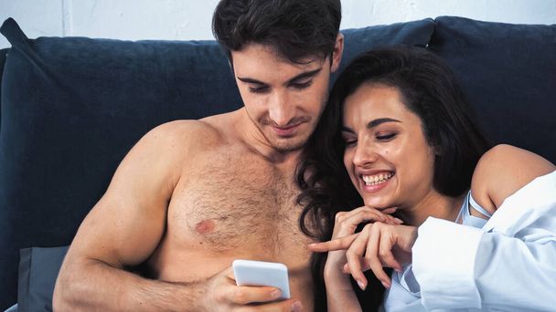 happy woman pointing with finger at smartphone near shirtless boyfriend in bedroom - Фото, зображення