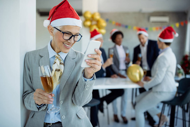 Boldog, érett üzletasszony SMS-ezik mobilon, miközben pezsgőt iszik szilveszterkor az irodában. A kollégái a háttérben vannak..  - Fotó, kép