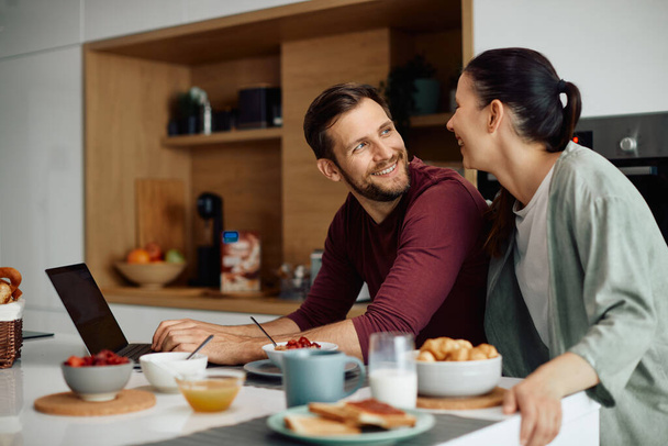 Young happy man communicating with his wife while using laptop during their breakfast at home. - Photo, image