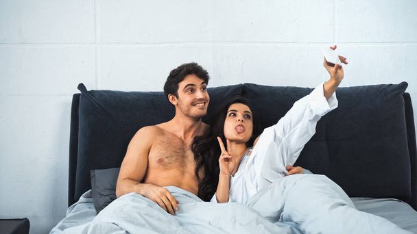 young woman sticking out tongue while taking selfie with cheerful and shirtless man in bed - Foto, Imagen