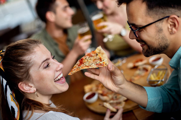 Щасливий чоловік годує свою дівчину піцою, обідаючи з друзями в пабі
.  - Фото, зображення
