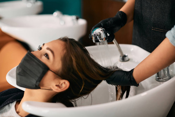 Молодая женщина носит защитную маску для лица во время мытья волос в парикмахерской. - Фото, изображение