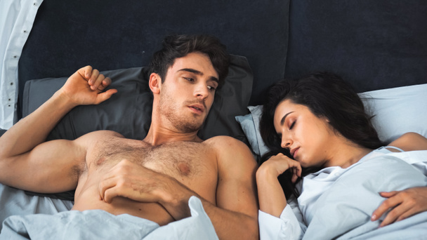 awakened shirtless man looking at brunette woman with closed eyes sleeping in bed - Fotó, kép