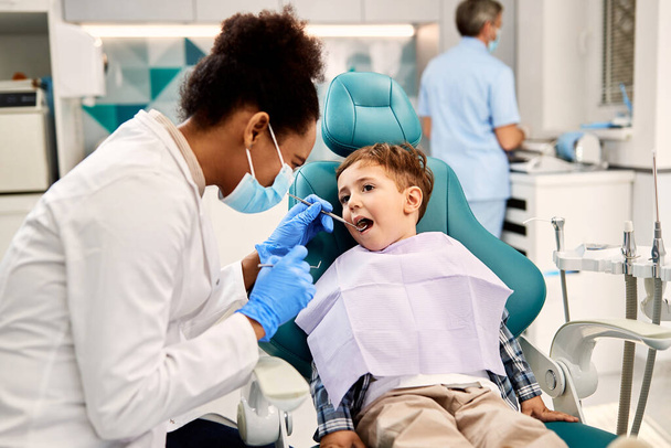 Чорна жінка-стоматолог вивчає зуби маленького хлопчика під час стоматологічної процедури в кабінеті стоматолога. Фокус на хлопчику
. - Фото, зображення