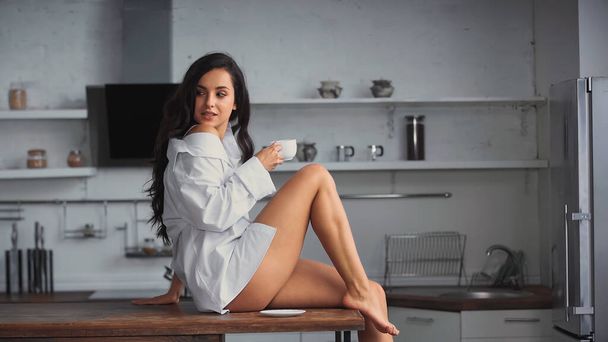barefoot young woman sitting on kitchen table and holding cup of coffee  - Foto, Imagem