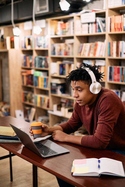 Young black student using laptop while drinking coffee and studying at university library. - 写真・画像