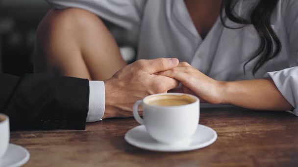 cropped view of man and woman holding hands near cup of coffee on kitchen table - Foto, imagen