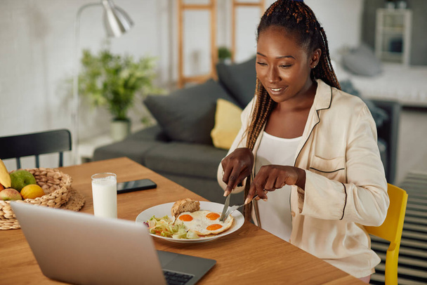 Young African American woman surfing the net on laptop while having breakfast at dining table. - Foto, Imagem