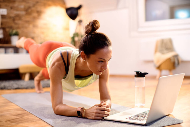 Dedicated sportswoman working out while following online exercise class over laptop at home. - Fotografie, Obrázek