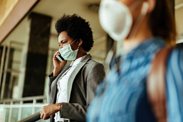 Чорна бізнес-леді носить маску для обличчя як захист від вірусів і розмовляє на мобільному телефоні під час ходьби в терміналі аеропорту
.  - Фото, зображення