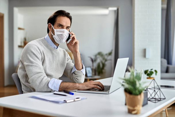 Young businessman wearing protective face mask while working on laptop and talking on mobile phone at home office.  - Photo, Image