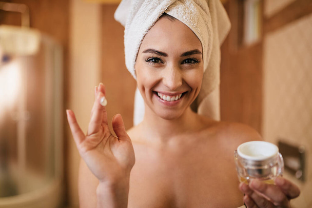 Beautiful woman using face cream and looking at camera in the bathroom. - Photo, image