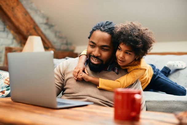 Small African American girl embracing her father while surfing the net on laptop together at home. - Foto, immagini