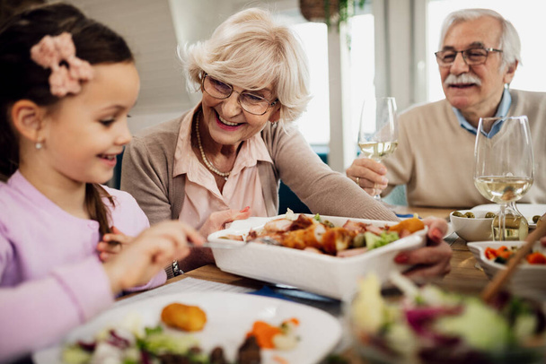 Happy grandparents and granddaughter having lunch at dining table. Focus is on senior woman offering food to a little girl. - Foto, afbeelding
