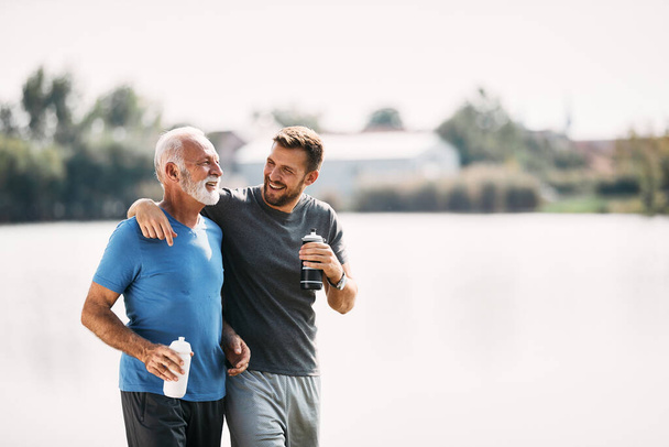 Senior athlete and his adult son talking and drinking water after running by the river in nature. Copy space. - Foto, Bild