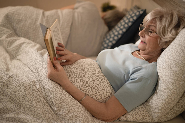Senior woman relaxing while reading a book in bed in the evening. Her husband is sleeping in the background.  - Foto, Imagem