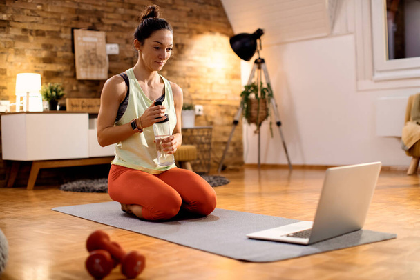 Smiling athletic woman refreshing herself with water while having online sports training over laptop from home. - Foto, Imagem